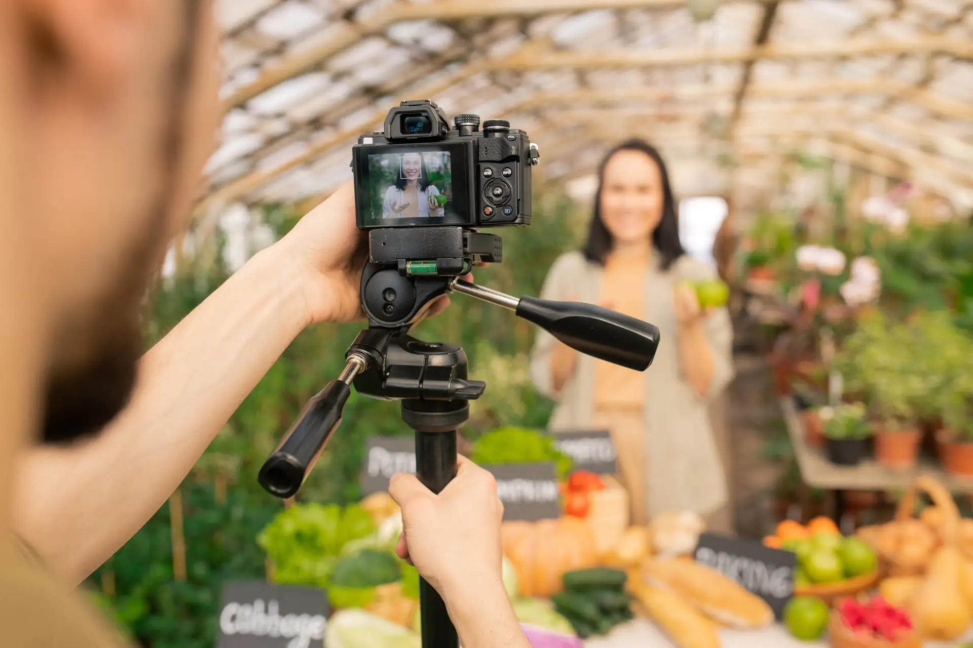 image taken from perspective of someone behind a camera, woman is being filmed, but she is blurred out, focus is on dlsr camera. The woman is in a greenhouse.